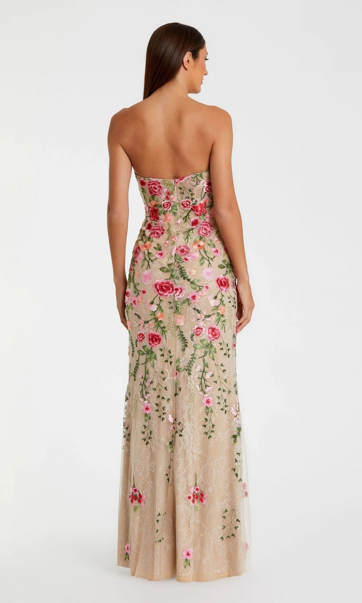 A woman faces away, wearing the Mac Duggal 68894 long formal dress—a strapless, form-fitting beige gown featuring pink and green floral embroidery and delicate lace details—set against a plain white background.